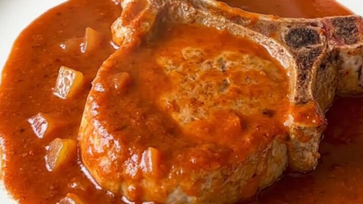 Tender pork chops smothered in vibrant red enchilada sauce with soft onions, served on a white plate.