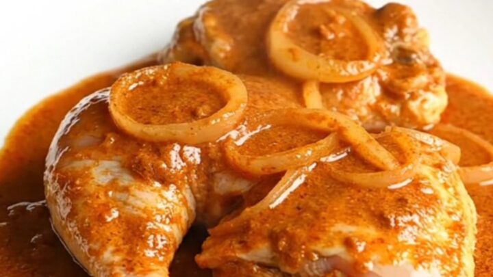 A serving spoon lifting a tender, bone-in chicken thigh coated in a rich, creamy orange paprika sauce from a slow cooker.