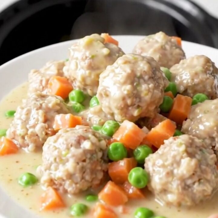 A close-up view of creamy chicken pot pie meatballs in a slow cooker, garnished with peas and carrots.