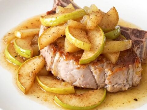 A close-up of a tender, bone-in pork chop topped with warm, spiced apple pie filling on a white plate.