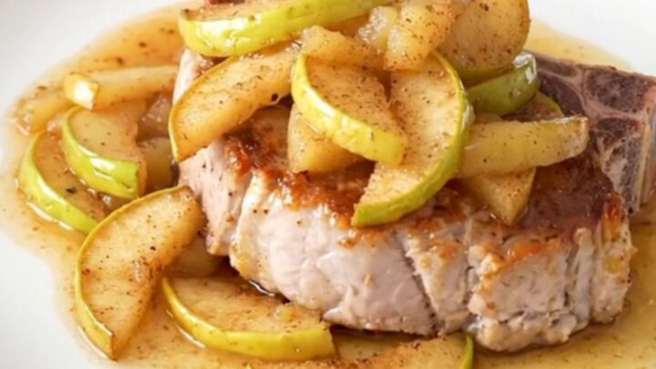 A close-up of a tender, bone-in pork chop topped with warm, spiced apple pie filling on a white plate.