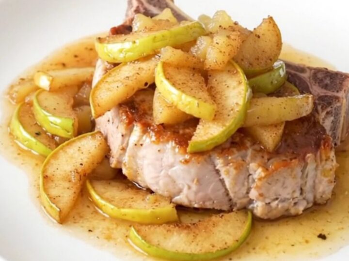 A close-up of a tender, bone-in pork chop topped with warm, spiced apple pie filling on a white plate.