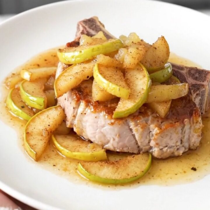 A close-up of a tender, bone-in pork chop topped with warm, spiced apple pie filling on a white plate.