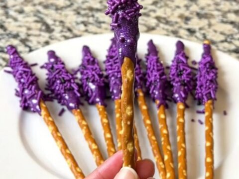A dozen purple-dipped witch wand pretzel rods arranged neatly on a dark baking sheet.