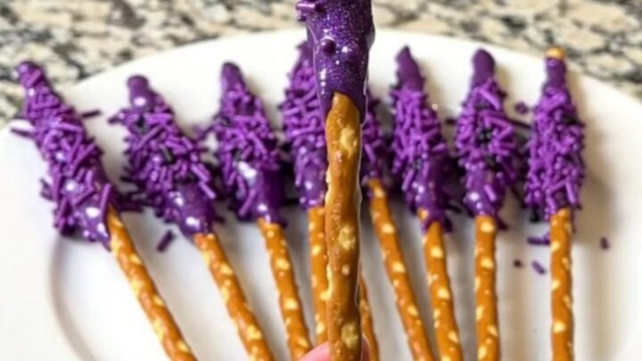A dozen purple-dipped witch wand pretzel rods arranged neatly on a dark baking sheet.