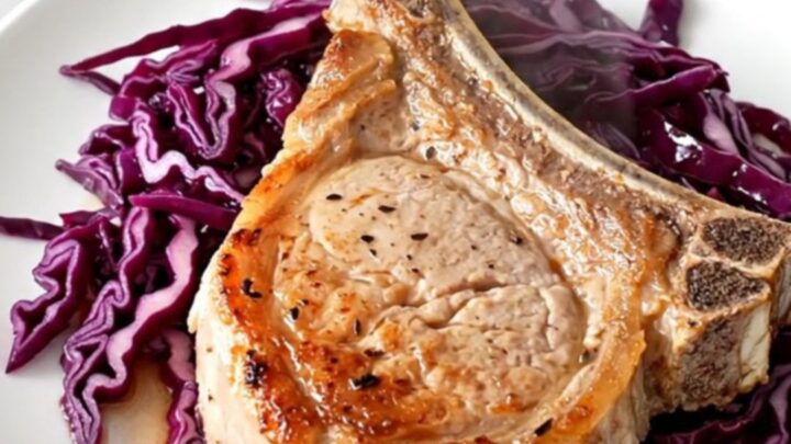 A close-up of a tender bone-in pork chop served over a bed of deep purple braised cabbage and sliced apples.