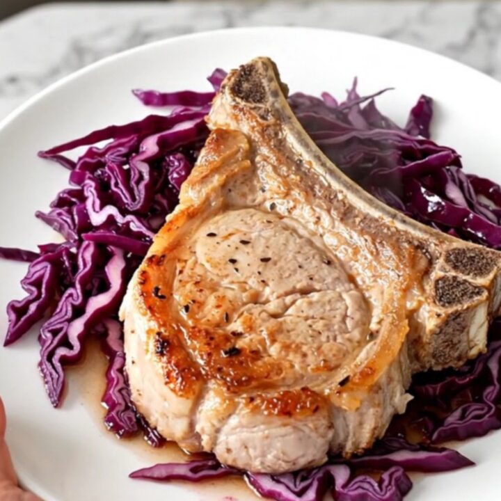 A close-up of a tender bone-in pork chop served over a bed of deep purple braised cabbage and sliced apples.