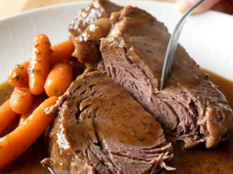 A rustic slow cooker filled with a tender, cooked chuck roast and bright orange baby carrots in a rich brown herb gravy.