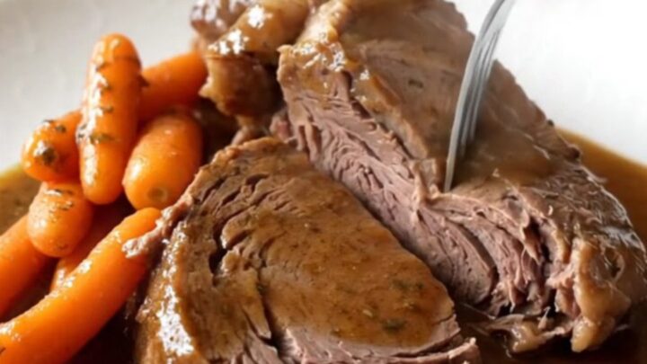 A rustic slow cooker filled with a tender, cooked chuck roast and bright orange baby carrots in a rich brown herb gravy.