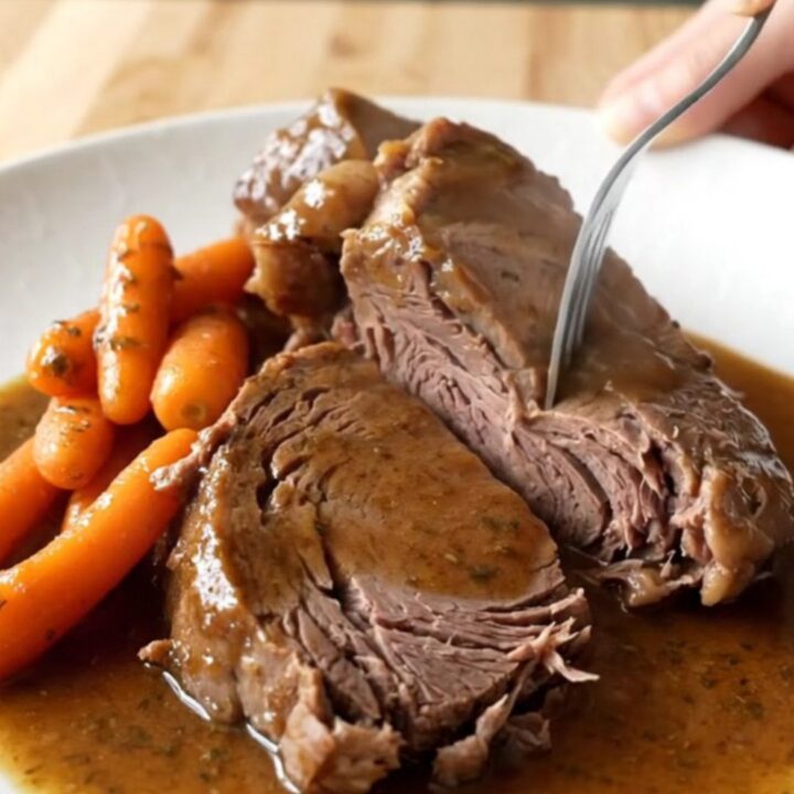 A rustic slow cooker filled with a tender, cooked chuck roast and bright orange baby carrots in a rich brown herb gravy.