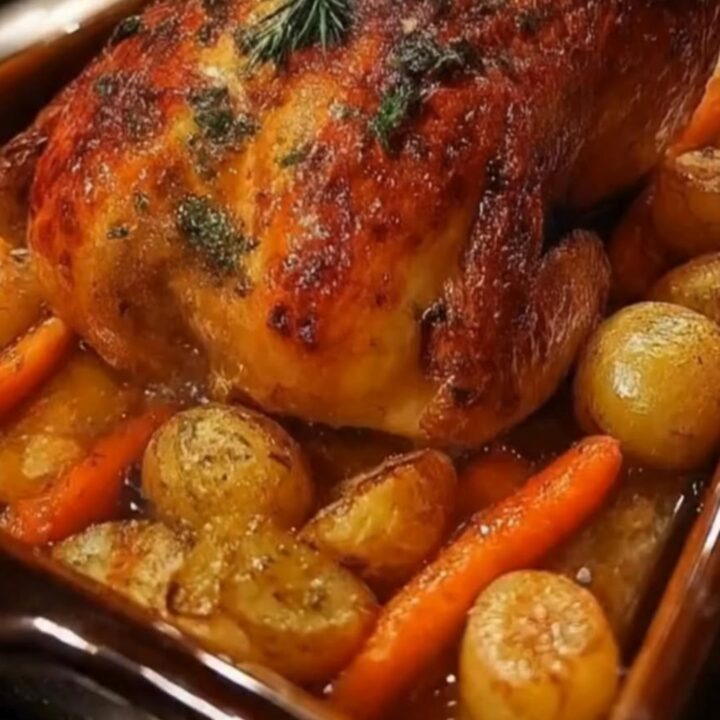A whole golden roasted herb chicken nestled with carrots and potatoes in a brown ceramic pan.