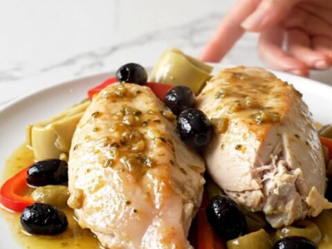 A close-up of tender, slow-cooked chicken breasts topped with artichokes, red peppers, and olives on a white plate.