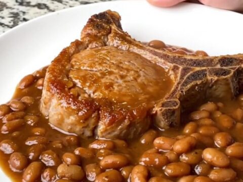 A serving of tender bone-in pork chop covered in rich BBQ baked beans on a white plate.