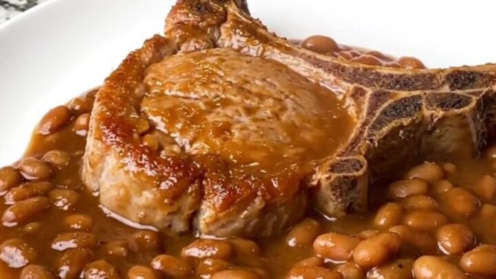A serving of tender bone-in pork chop covered in rich BBQ baked beans on a white plate.