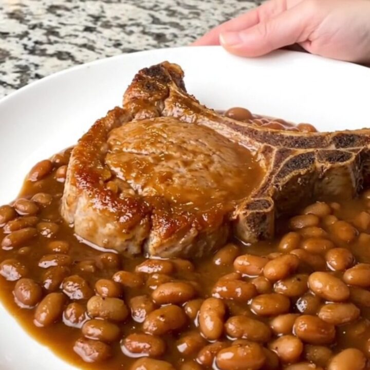 A serving of tender bone-in pork chop covered in rich BBQ baked beans on a white plate.