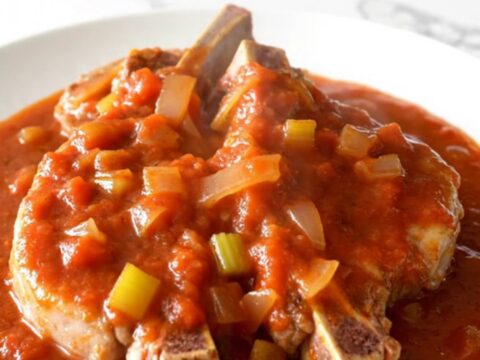 Bone-in pork chops smothered in a thick, rich tomato gravy with visible onions and celery, served on a white plate.