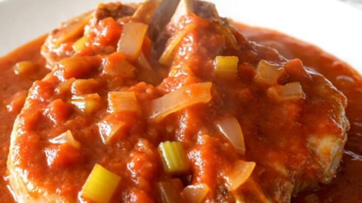 Bone-in pork chops smothered in a thick, rich tomato gravy with visible onions and celery, served on a white plate.