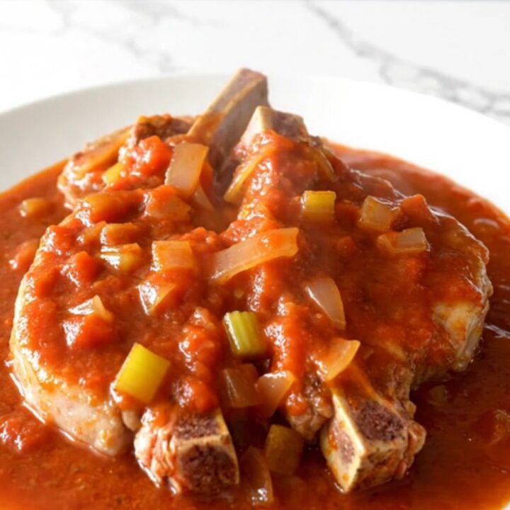Bone-in pork chops smothered in a thick, rich tomato gravy with visible onions and celery, served on a white plate.