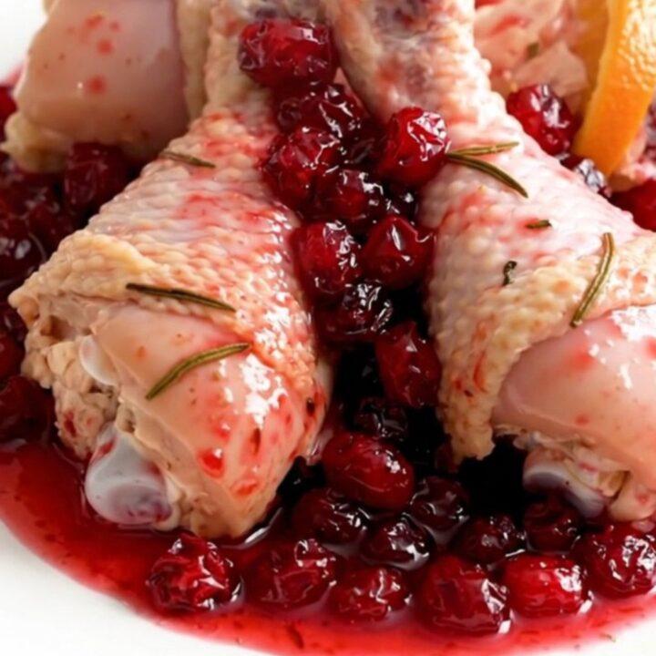 A close-up of slow-cooked chicken drumsticks coated in a glossy, vibrant cranberry and orange sauce on a white plate.