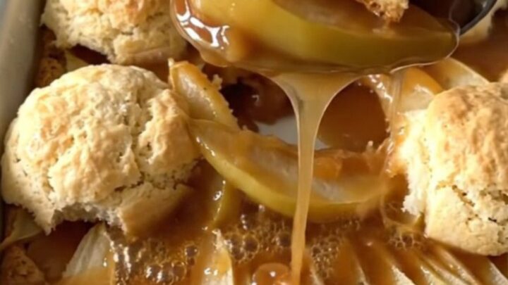 A warm, golden brown pear cobbler in an enameled baker with a serving spoon lifting a portion.