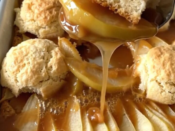 A warm, golden brown pear cobbler in an enameled baker with a serving spoon lifting a portion.