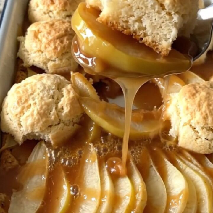 A warm, golden brown pear cobbler in an enameled baker with a serving spoon lifting a portion.