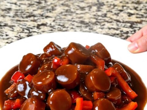 A close-up of slow-cooked smoked sausage coins glazed in a thick, dark General Tso sauce with red bell peppers.