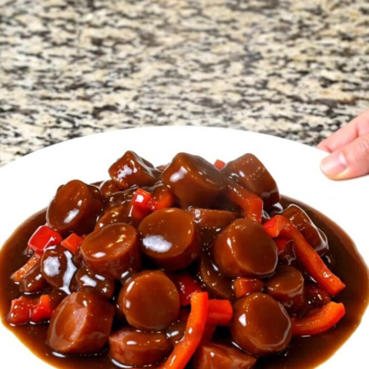 A close-up of slow-cooked smoked sausage coins glazed in a thick, dark General Tso sauce with red bell peppers.