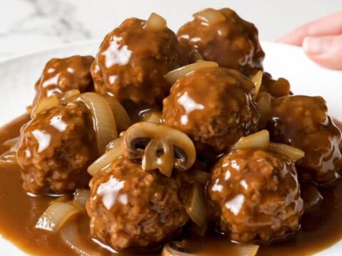 A close-up of slow cooker Salisbury steak meatballs served on a white plate with rich gravy and soft onions.