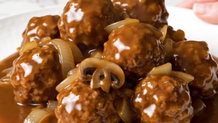 A close-up of slow cooker Salisbury steak meatballs served on a white plate with rich gravy and soft onions.