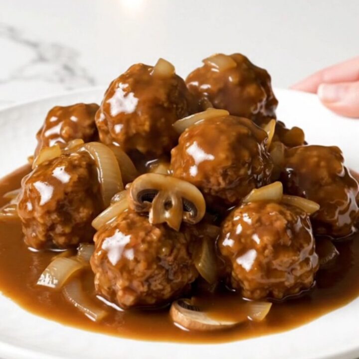 A close-up of slow cooker Salisbury steak meatballs served on a white plate with rich gravy and soft onions.