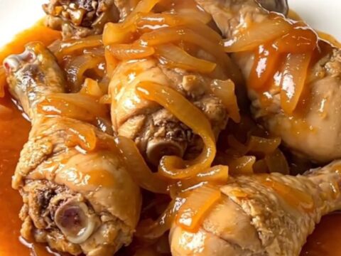 A top-down view of skinless chicken drumsticks arranged in a ring inside a black slow cooker with onions and sauce.