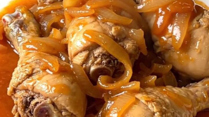A top-down view of skinless chicken drumsticks arranged in a ring inside a black slow cooker with onions and sauce.