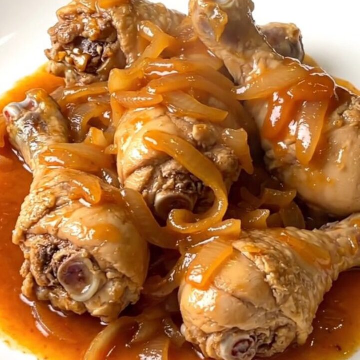 A top-down view of skinless chicken drumsticks arranged in a ring inside a black slow cooker with onions and sauce.