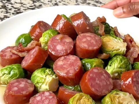 A top-down view of a black slow cooker filled with sliced kielbasa, halved Brussels sprouts, and chopped bacon.