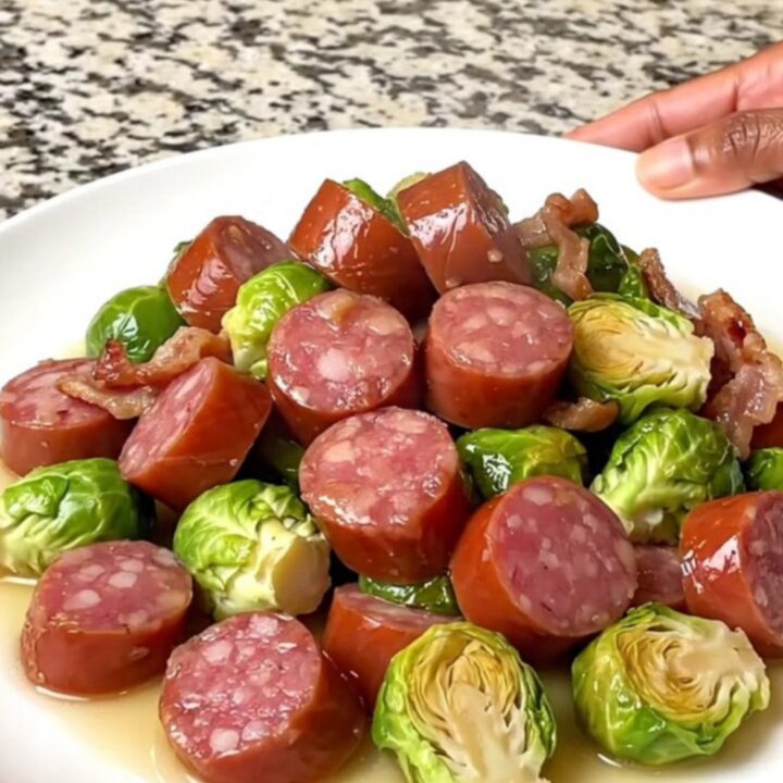 A top-down view of a black slow cooker filled with sliced kielbasa, halved Brussels sprouts, and chopped bacon.