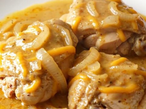 A close-up of tender chicken thighs smothered in a creamy, thick cheddar sauce with soft onion slices.