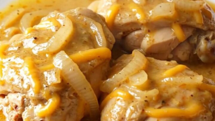 A close-up of tender chicken thighs smothered in a creamy, thick cheddar sauce with soft onion slices.
