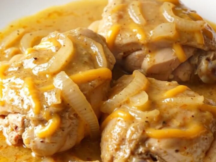 A close-up of tender chicken thighs smothered in a creamy, thick cheddar sauce with soft onion slices.