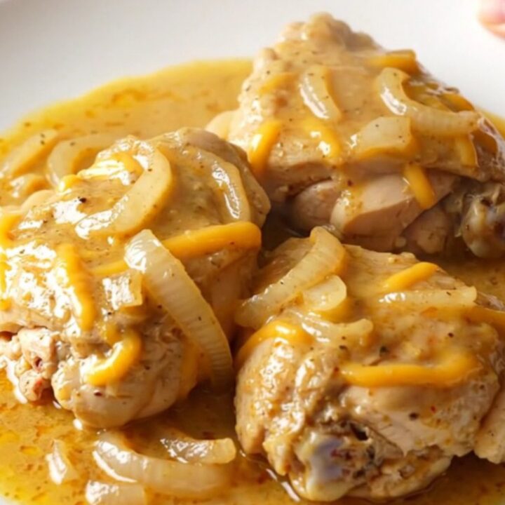A close-up of tender chicken thighs smothered in a creamy, thick cheddar sauce with soft onion slices.