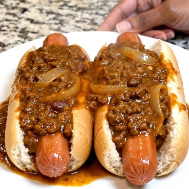 A slow cooker filled with plump hot dogs covered in a thick, hearty chili sauce on a granite countertop.