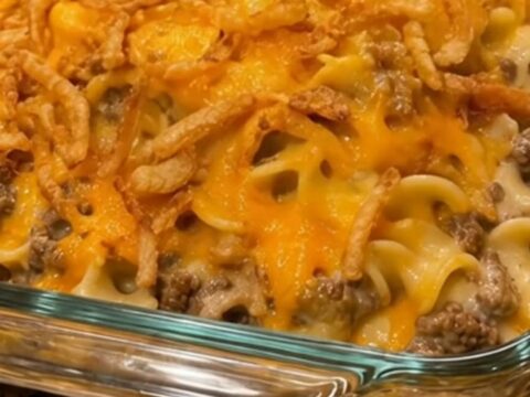 A steaming, golden cheesy beef and noodle casserole in a glass dish topped with crispy fried onions.