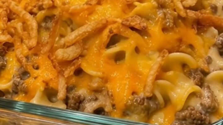 A steaming, golden cheesy beef and noodle casserole in a glass dish topped with crispy fried onions.