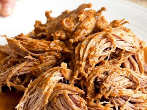 A close-up of tender, shredded pork barbacoa in a slow cooker, showing thick red juices and steam.