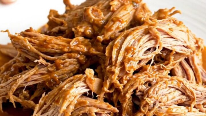 A close-up of tender, shredded pork barbacoa in a slow cooker, showing thick red juices and steam.
