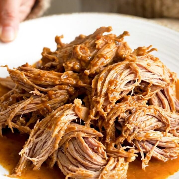 A close-up of tender, shredded pork barbacoa in a slow cooker, showing thick red juices and steam.