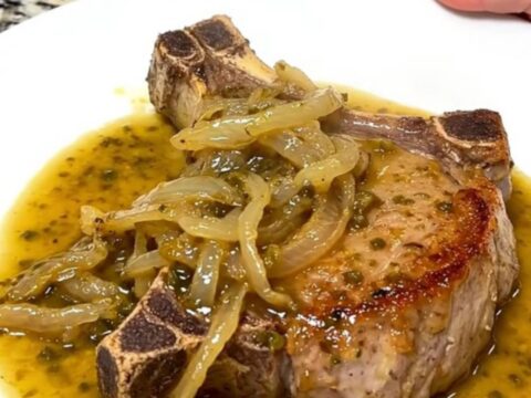 A close-up of tender, slow-cooked pork chops covered in a rich chili lime sauce and soft red onions on a white plate.