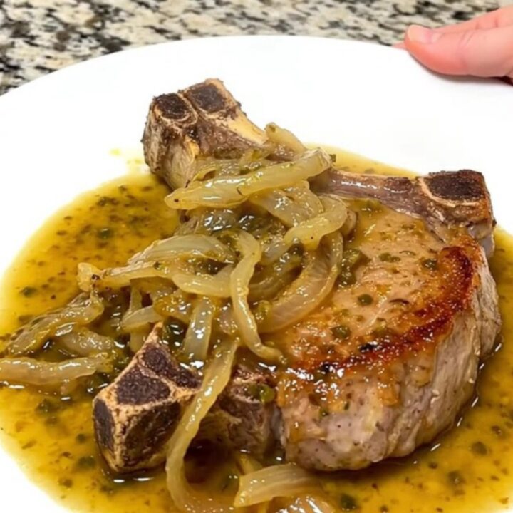 A close-up of tender, slow-cooked pork chops covered in a rich chili lime sauce and soft red onions on a white plate.