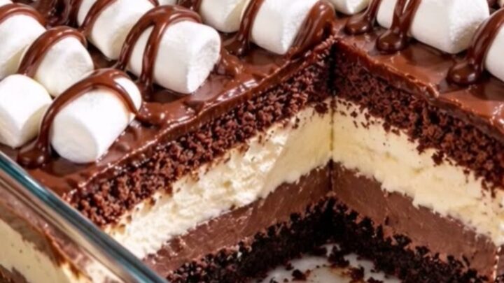 A glass casserole dish filled with layered crumb crust, pudding, cream, cake, and marshmallows drizzled with ganache.