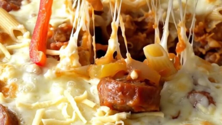 A top-down view of a ceramic baking dish filled with raw penne, sausage coins, bell peppers, and onions.